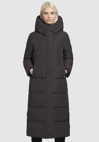 Zwarte pufferjas met een lange silhouette, grote capuchon en belangrijke kenmerken zoals ritszakken en een gestikte afwerking. Glad textuur en strakke afwerking.