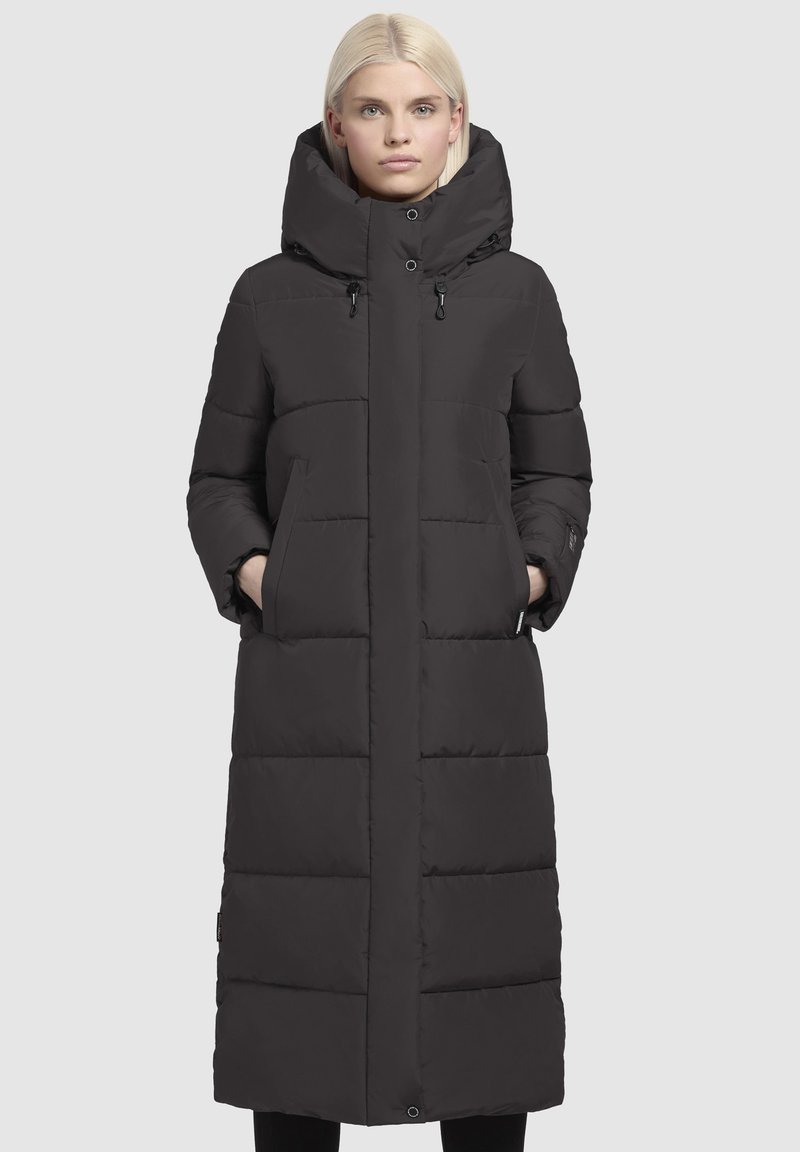 Zwarte pufferjas met een lange silhouette, grote capuchon en belangrijke kenmerken zoals ritszakken en een gestikte afwerking. Glad textuur en strakke afwerking.