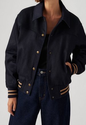 Kvinde iført en sort bomberjakke med knapper og beige stribede ribmanchetter og kant, kombineret med mørkeblå, højtaljede jeans.