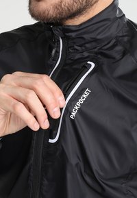 Veste coupe-vent noire avec poche poitrine zippée et accents réfléchissants. Texture lisse avec un col montant et des réglages ajustables.