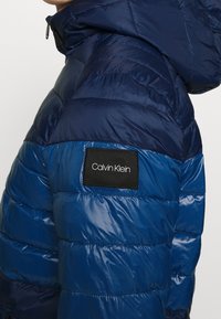 Vadderad blå jacka med en glänsande yta, med huva, horisontella quiltade sektioner och ett svart märke med "Calvin Klein"-logotypen på axeln.