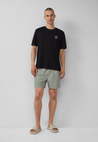 Zwart katoenen T-shirt met een klein logo, gecombineerd met lichtgroene shorts en beige instapschoenen. Eenvoudig, casual ontwerp met een ontspannen pasvorm.