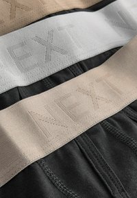Bavlněné spodní prádlo s třemi elastickými pásky v světle béžové, šedé a černé barvě, každý s vyraženým nápisem "NEXT" ve stejných barvách.