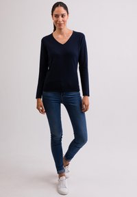 Námořnický v-neck svetr vyrobený z měkkého materiálu, zkombinovaný s úzkými modrými džíny a bílými teniskami. Model stojí uvolněně na čistém pozadí.