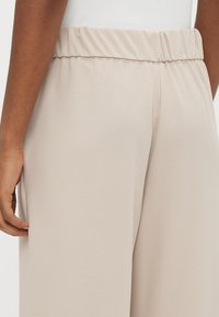 Μπεζ παντελόνι με ελαστική μέση, με λεία υφή και ίσια εφαρμογή, εμ φανίζεται εν μέρει με ένα χέρι τοποθετημένο στο πλάι.