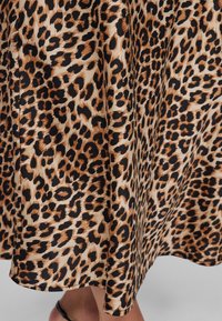 Leopárdmintás szoknya anyaga fekete és barna foltokkal bézs háttéren, részben elfedve egy személy alsó lábait és fekete szandált.