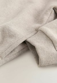 Tessuto di maglione grigio chiaro con superficie testurizzata. Presenta polsini a costine e una finitura pulita, mettendo in evidenza un materiale morbido e accogliente.