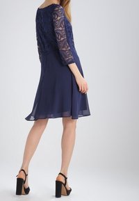 Námořnicky modré šaty s vyšitým krajkovým živůtkem, rukávy tři čtvrtě, a splývavou hedvábnou sukní; doplněné černými sandály na vysokém podpatku.