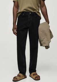 Vaqueros de mezclilla negros con un ajuste relajado, llevados con una camisa de manga corta verde. Una chaqueta beige se sostiene en la mano izquierda; sandalias marrones en los pies.