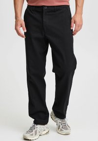 Pantalones negros de tela elástica, con corte recto, cierre de botones y bolsillos laterales, combinados con zapatillas grises y blancas.