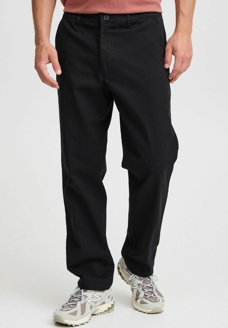 Pantalones negros de tela elástica, con corte recto, cierre de botones y bolsillos laterales, combinados con zapatillas grises y blancas.