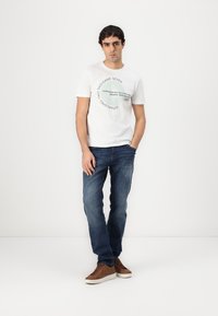 Hvit t-skjorte med sirkelformet grafisk tekst, blå jeans og brune joggesko. Jeansene har en lett falming og rett snitt.