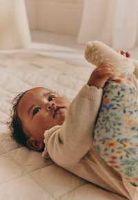 Baby ligger på en polstret tæppe, holder på et ben, iført en cremet sweater og bukser med blomsterprint i et blødt belyst rum.