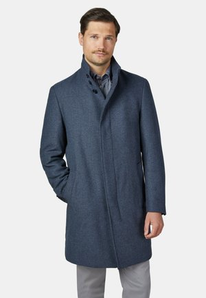 Mann trägt einen blauen wollenen Mantel mit Knöpfen und graue Hose, mit einer Hand in der Tasche, vor einem einfarbigen hellen Hintergrund stehend.