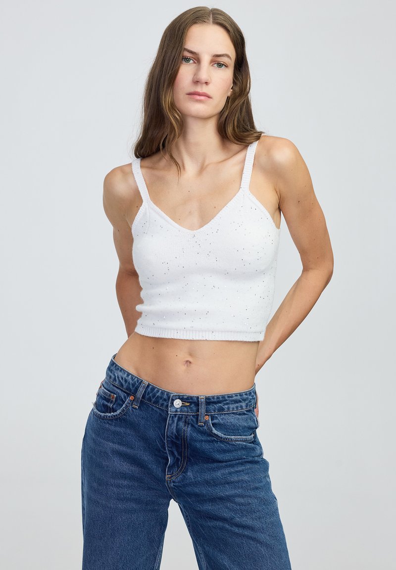 Biały krótki top na ramiączkach z dekoltem w kształcie litery V, cienkimi paskami, subtelnymi plamkami i prążkowanym dołem. W zestawie z niebieskimi dżinsami z wysokim stanem.