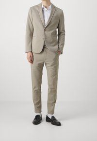 Uomo in completo beige con camicia bianca, mocassini neri e calzini bianchi, in piedi con una mano in tasca su sfondo grigio.