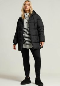 Doudoune noire avec capuche, présentant un design matelassé. Superposée à un pull gris rayé et un pantalon noir, associée à des bottines noires.