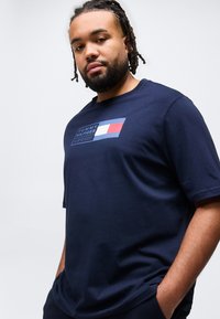 Άνδρας με πλεξούδες και γένια, φορώντας μπλε ναυτικό μπλουζάκι Tommy Hilfiger με λογότυπο, στέκεται με τα χέρια στις τσέπες μπροστά σε απλό φόντο.