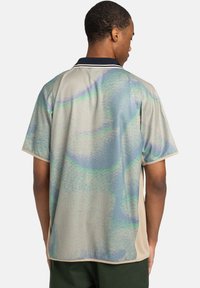 Kurzarm-Polohemd in hellbeige mit einem wirbelnden grünen und blauen Muster. Verfügt über einen marineblauen Kragen mit kontrastierenden Besätzen.