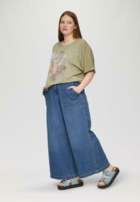 Oliwkowy T-shirt z grafiką, zestawiony z szerokimi niebieskimi dżinsami i jasnoniebieskimi sandałami z podwójnymi klamrami.