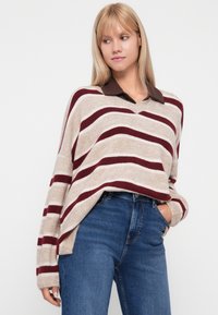 Overdimensie sweater met een V-hals, beige basis en bordeauxrode strepen. Bruin kol onderkleding zichtbaar. Gecombineerd met blauwe spijkerbroeken.