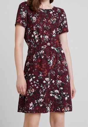 Femme portant une robe à fleurs bordeaux à manches courtes avec une taille élastique, debout devant un fond uni gris clair.