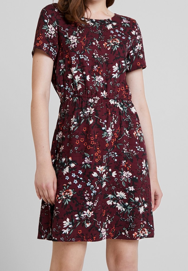 Femme portant une robe à fleurs bordeaux à manches courtes avec une taille élastique, debout devant un fond uni gris clair.