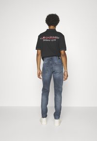 Camisa negra de manga corta con el texto rojo y blanco "Gucci Milano 1978" en la parte de atrás, combinada con jeans ajustados azules y zapatillas blancas.
