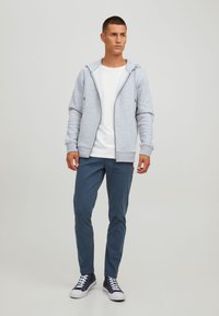 Lys grå lynlås hættetrøje med hætte, forlommer, ribbede manchetter; parret med blå bukser og mørke sneakers, stående på en neutral baggrund.