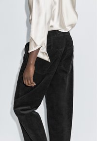 Zwarte corduroy broek met een gestructureerde afwerking, voorzien van achterzakken en een losse pasvorm. Een crèmekleurige satijnen blouse is gedeeltelijk zichtbaar.