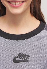 Grijs mesh stof T-shirt met een zwarte geribbelde halslijn en een zwart Nike-logo op de borst. Close-up weergave met focus op de kraag en logodetails.