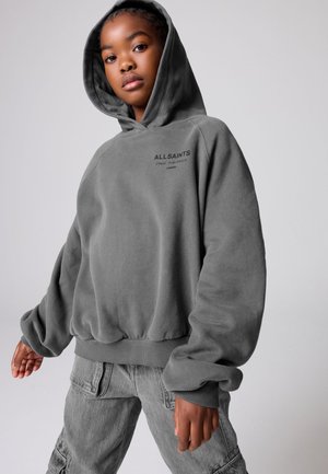Junge Person trägt einen lockeren grauen Kapuzenpullover mit dem Text „ALLSAINTS LONDON“ und graue Cargohosen, posiert vor einem einfarbigen Hintergrund.