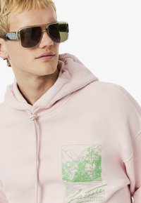 Sudadera rosa con cordón y diseño gráfico verde que muestra montañas y árboles. Llevada con gafas de sol de montura transparente y lentes oscuros.