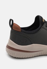 Schwarzer Sportschuh mit strukturiertem Obermaterial, braunem Stoffakzent an der Ferse und einer hellen Gummisohle mit Wellenmuster. Logo an der Seite.