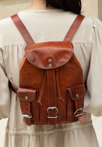 Brown leather backpack with front flap, buckle straps, and side pockets worn over beige long-sleeve garment.