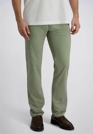 Man draagt lichtgroene rechte broek, wit overhemd en donkerbruine nette schoenen, staand tegen een effen achtergrond.