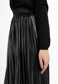 Sorte plisseret nederdel lavet af skinnende materiale, med en bred elastisk talje. Stoffet har en glat tekstur med fremtrædende lodrette folder.