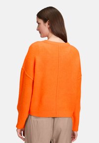 Kobieta z długimi brązowymi włosami, ubrana w jasnopomarańczowy, puszysty sweter i beżowe spodnie, widziana od tyłu na białym tle.