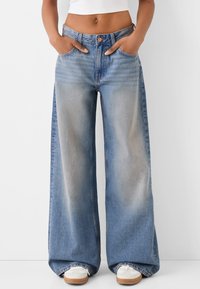 Henkilö, joka pitää käsiään taskuissa valkoisen taustan edessä, pukeutuneena vaaleansinisiin leveisiin farkkuihin ja valkoisiin tennareihin.