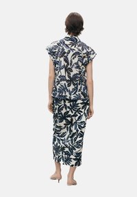 Personne debout de dos, portant un haut sans manches à motif de feuilles blanches et bleu marine et une jupe midi assortie, avec des talons ouverts beiges.