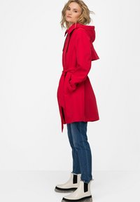 Červený voděodolný trench coat s kapucí, opaskem v pase a bočními kapsami, spárovaný s modrými džínami a krémovými kotníkovými botami.