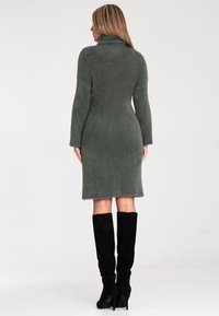 Strickkleid in gedämpftem Grün mit hohem Kragen, langen Ärmeln und taillierter Silhouette, kombiniert mit knielangen schwarzen Wildlederstiefeln.