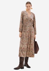 Květinové dlouhé šaty s dlouhým rukávem v béžové barvě s fialovými a zelenými vzory, stahovací pas, v kombinaci s hnědými kozačkami po kolena a tmavě hnědou taškou.