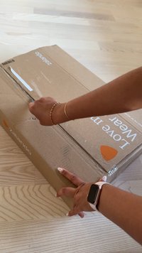 Brown cardboard box with Zalando branding, partially opened with a hand holding the edge. The surface is flat with a wooden floor beneath.