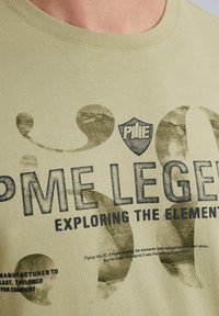 Oliwkowa koszulka z krótkim rękawem, z ciemnym nadrukiem tekstu i wzorów. Tekst zawiera "PME LEGEND" oraz "EXPLORING THE ELEMENTS."