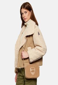 Giacca in shearling beige e crema con collo ampio, indossata sopra un leggero dolcevita. Il modello tiene una piccola borsa a tracolla beige con dettagli testurizzati.