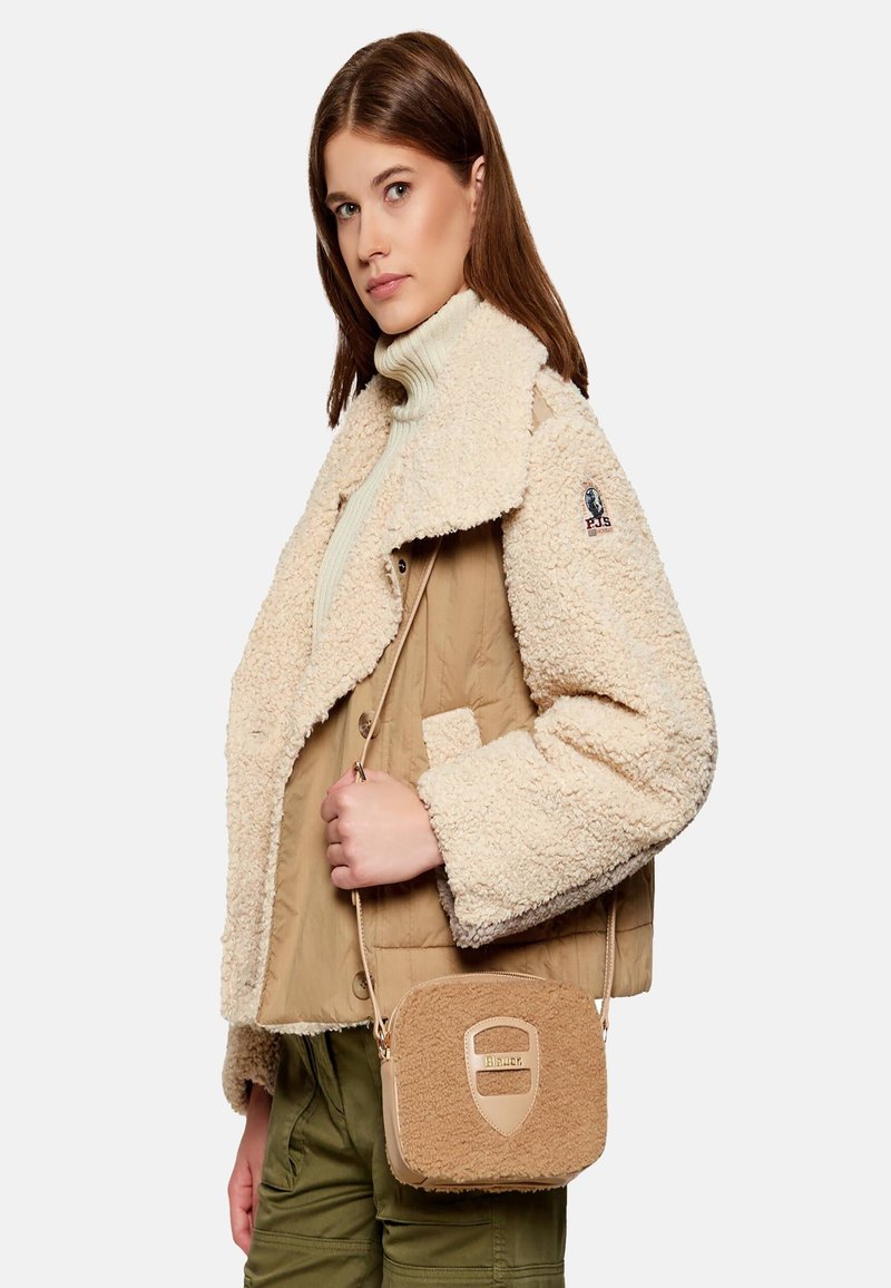 Giacca in shearling beige e crema con collo ampio, indossata sopra un leggero dolcevita. Il modello tiene una piccola borsa a tracolla beige con dettagli testurizzati.