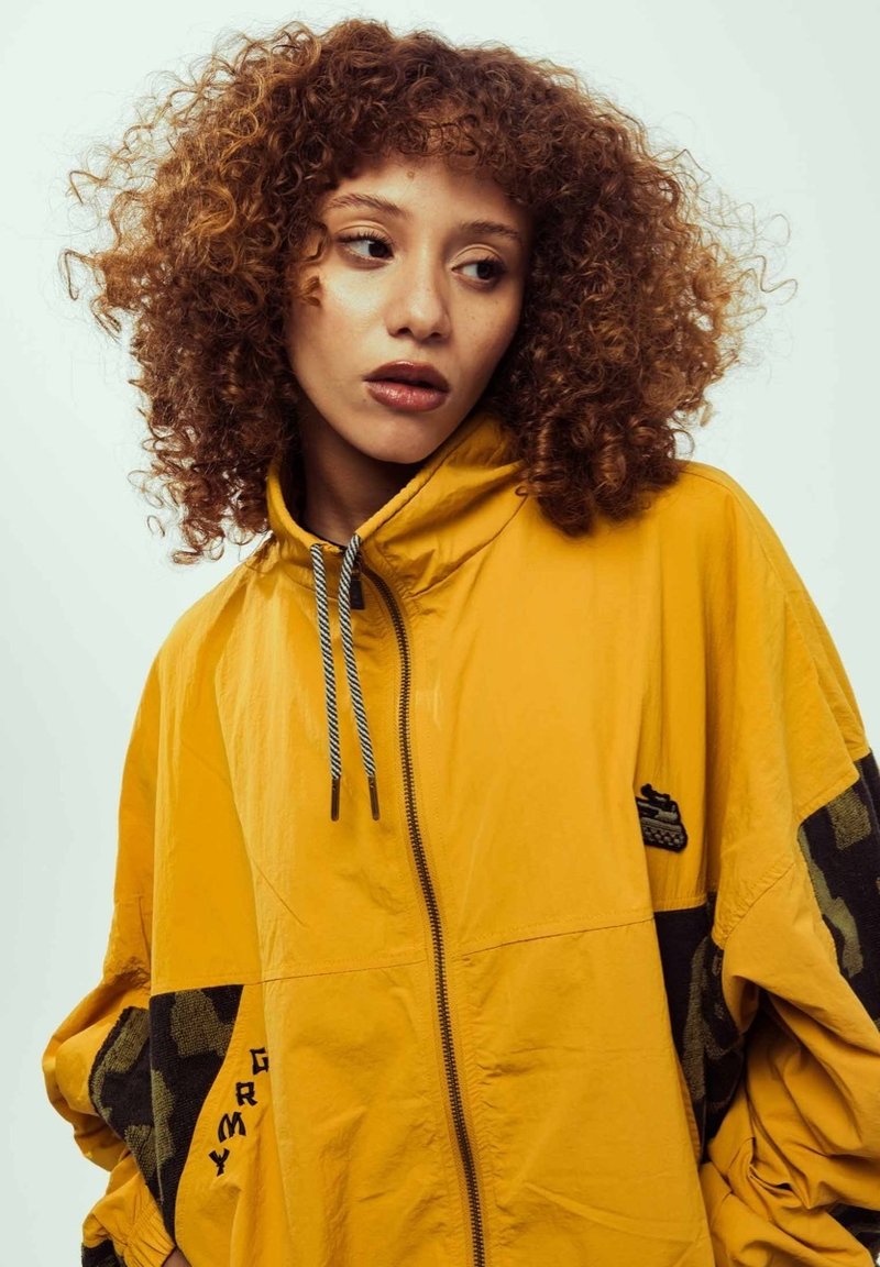 Chaqueta amarilla con cremallera y capucha, con detalles en negro y mangas de camuflaje. Fabricada en tela ligera con un corte relajado y detalles de cordón.