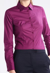 Blusa aderente a maniche lunghe di un profondo viola, realizzata in tessuto morbido. Presenta un colletto classico e una fila di bottoni lungo il davanti.