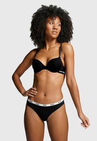 Schwarzes Bralette mit verstellbaren Trägern und gepolsterten Cups, kombiniert mit einem schwarzen Tanga mit einem weißen, gebrandeten Bund, glatter Stofftextur.